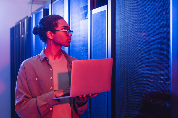 Африканский американский техник с ноутбуком смотрит на серверы в центре обработки данных в неоновом свете - Фото, изображение