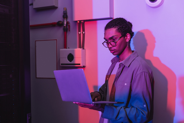 young african american programmer in eyeglasses using laptop in data center in neon light - Photo, Image