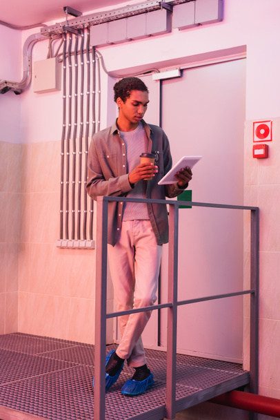 joven ingeniero americano afican mirando tableta digital mientras está de pie con bebida para llevar en el centro de datos - Foto, Imagen