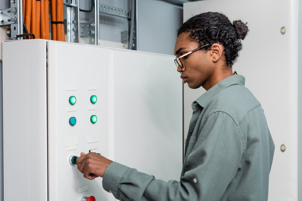 joven técnico afroamericano pulsando botón en centralita mientras trabaja en centro de datos - Foto, imagen
