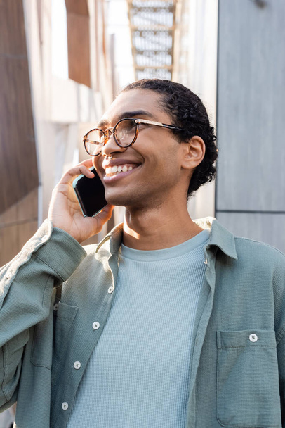 joyful african american man in grey shirt and eyeglasses talking on smartphone on urban street - Photo, Image