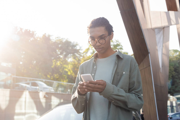 homme afro-américain à la mode dans la messagerie de lunettes sur téléphone mobile en plein air - Photo, image