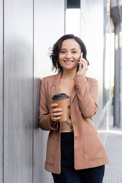 onnellinen nainen kahvia mennä katsomaan kameraa puhuessaan älypuhelimessa ulkona - Valokuva, kuva