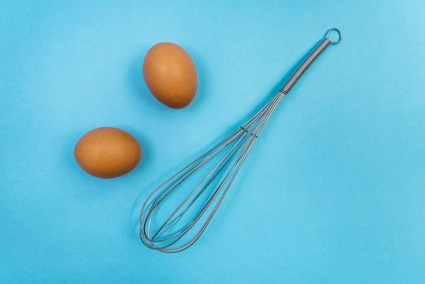 Two brown eggs and a whisk for whipping on a blue background. Natural products and kitchen equipment - Foto, Bild