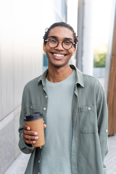 cheerful african american man in grey shirt and eyeglasses holding takeaway drink on city street - Photo, Image
