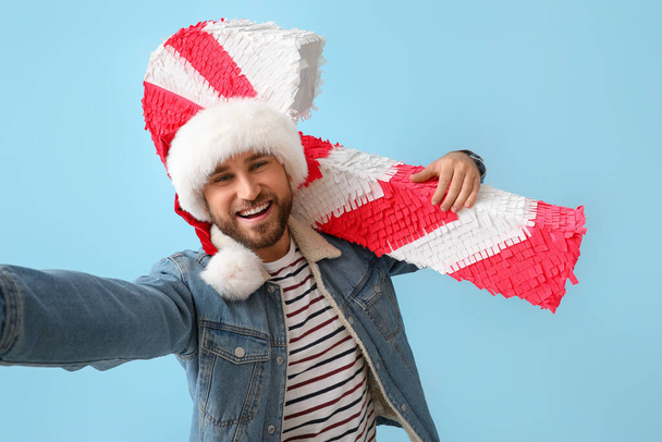 Hombre guapo en sombrero de Santa con piñata de bastón de caramelo tomando selfie sobre fondo azul - Foto, imagen