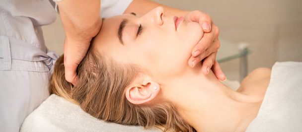 Kosmetolog rukou dělá masáž obličeje na čele a bradě mladé ženy v lázeňském salonu - Fotografie, Obrázek