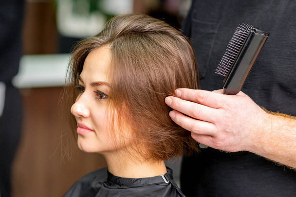 Мужской парикмахер работает над прической молодой кавказской брюнетки в парикмахерской - Фото, изображение