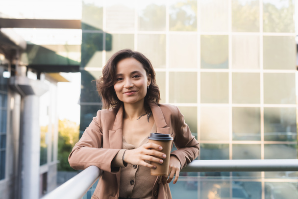 positive woman with takeaway drink looking at camera on balcony outdoors - Photo, Image
