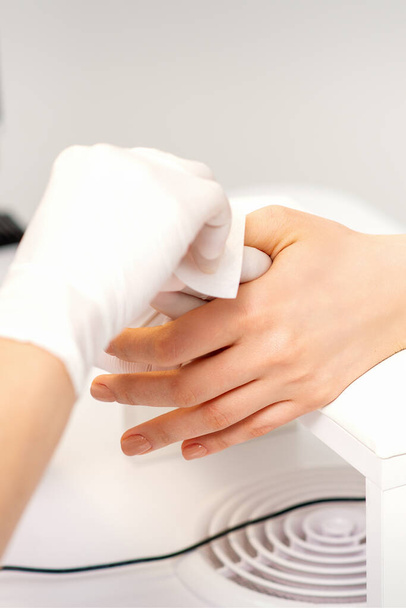 Las manos de una manicurista con guantes protectores blancos limpian las uñas femeninas con una servilleta de papel en el salón - Foto, Imagen
