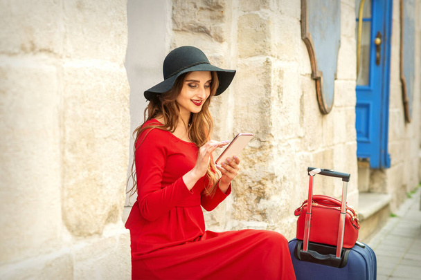 Una joven mujer blanca viajando en la chaqueta roja y sombrero negro sentado con un teléfono inteligente en la calle de la ciudad - Foto, imagen
