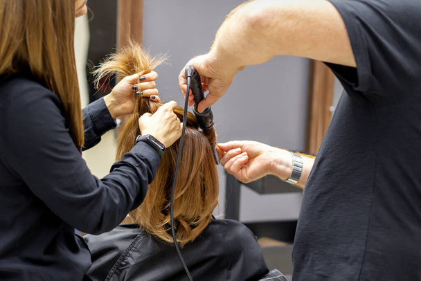 Η πίσω όψη των δύο κομμωτές είναι μπούκλες μαλλιά για μια νεαρή γυναίκα με ηλεκτρικό σίδερο μαλλιών σε ένα σαλόνι ομορφιάς - Φωτογραφία, εικόνα