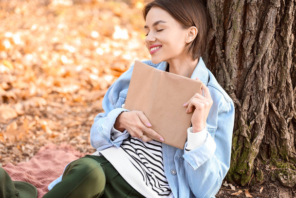 Felice donna sognante seduta vicino all'albero con libro - Foto, immagini