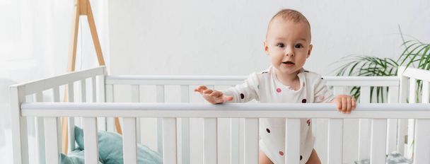 Baby im Strampler blickt in die Kamera, während es im Kinderbett steht, Banner - Foto, Bild