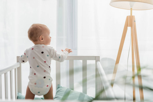 Rückansicht eines Kleinkindes im Babystrampler, das im Kinderbett steht - Foto, Bild