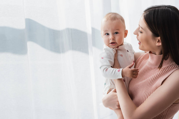 feliz morena mujer sosteniendo bebé niño cerca blanco cortina en casa - Foto, imagen