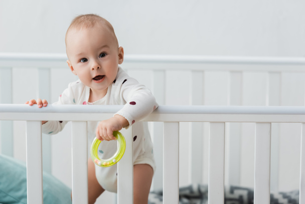 little boy holding rattle ring while standing in crib and looking at camera - Photo, Image