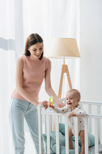 brunette woman smiling near little son standing in crib with rattle ring - Photo, Image