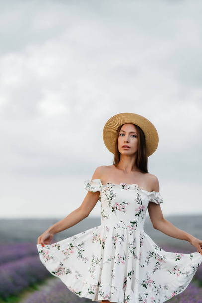 A young beautiful girl in a delicate dress and hat walks through a beautiful field of lavender and enjoys the aroma of flowers. Vacation and beautiful nature. - Fotó, kép