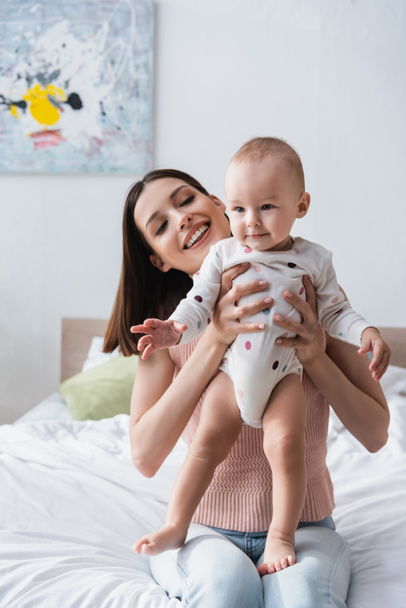 happy brunette woman holding toddler boy in romper while sitting on bed - Zdjęcie, obraz