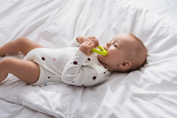 niño pequeño mordiendo juguete mientras está acostado en ropa de cama blanca en mameluco - Foto, imagen