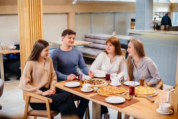 A group of young cheerful friends is sitting in a cafe talking and eating pizza. Lunch at the pizzeria. - Foto, Imagen