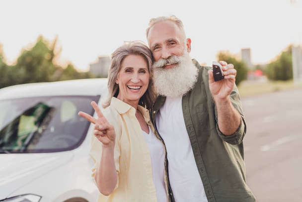 Photo of retired pensioner grey haired wife husband old people new car keys v-sign cool outside outdoors in city - Fotoğraf, Görsel