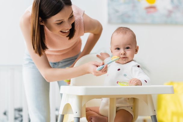 little boy in romper sitting in baby chair while happy mother feeding him from spoon - Photo, Image