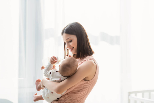 happy brunette woman feeding toddler from baby bottle while standing near white curtain - Photo, Image