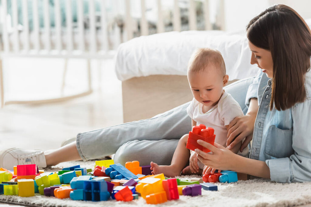 woman holding building block while playing with baby boy on floor in bedroom - Photo, Image