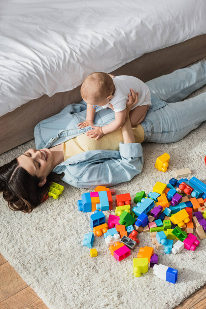 high angle view of happy woman lying on floor near building blocks while playing with toddler son  - Photo, Image