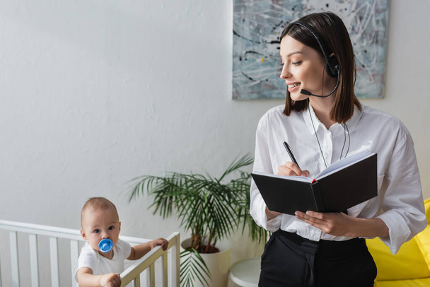 woman in headset writing in notebook while working at home near baby in crib - Foto, Imagem