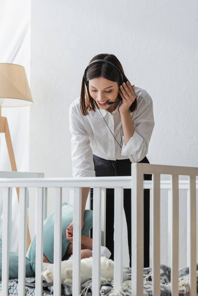 positive woman talking in headset near crib with toddler kid - Foto, Bild