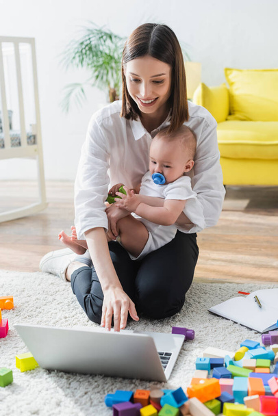 smiling woman using laptop while sitting on floor with son near building blocks - Foto, Bild