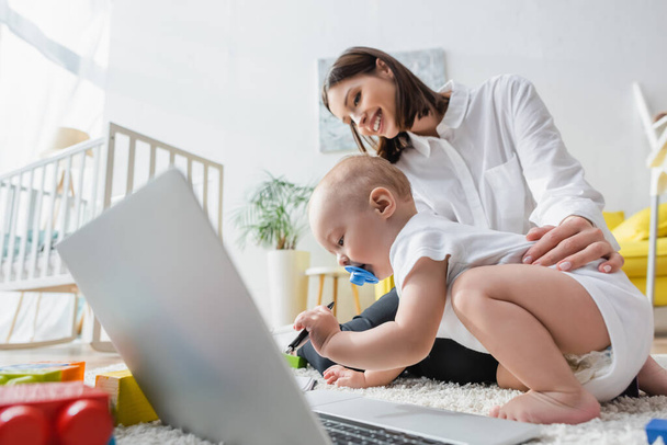 Kleinkind hält Stift in der Nähe von glücklicher Mutter und Laptop auf dem Boden - Foto, Bild