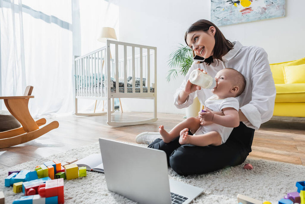 smiling woman talking on smartphone while feeding son on floor near laptop and building blocks - Foto, Bild