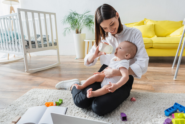 happy woman sitting on floor near laptop and feeding little son from baby bottle - Photo, Image