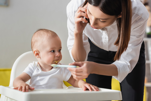 woman talking on mobile phone while feeding toddler son with puree - Photo, Image