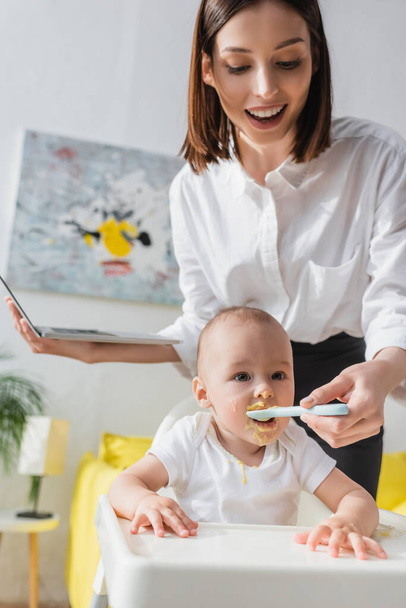 smiling woman holding laptop while feeding baby boy with puree at home - Valokuva, kuva