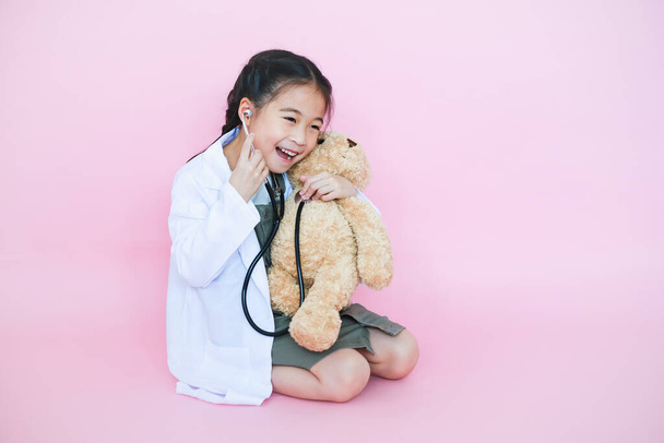 夢を見ている小さなアジアの子供の女の子は、ピンクの背景にテディベアと聴診器を使用して獣医師です - 写真・画像