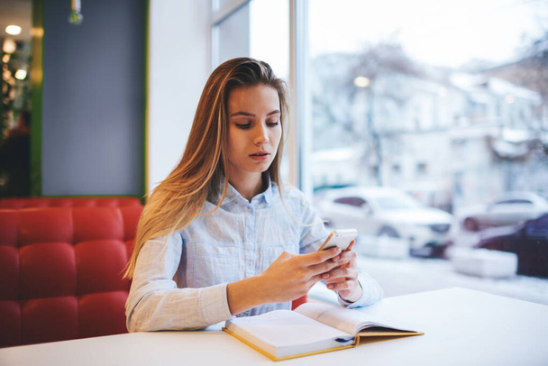 Young female student in casual outfit with long hair sitting at table with textbook and surfing smartphone while doing homework - Zdjęcie, obraz