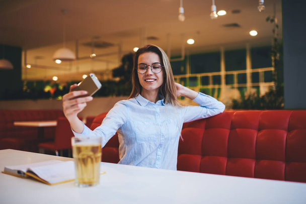 Positive female in casual clothes with toothy smile sitting on sofa leaning on hand and taking selfie on mobile phone while resting in light cafeteria - Fotó, kép