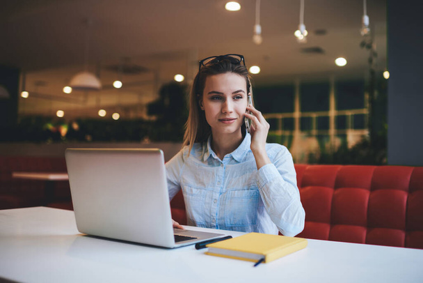 Calm female freelancer in casual clothes looking away while sitting at table in light workspace with laptop and using smartphone for calling - Foto, Bild