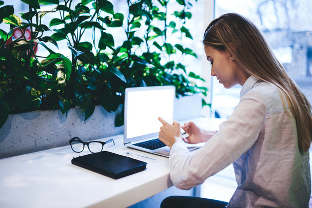 Side view of concentrated long haired female freelancer in white shirt messaging on smartphone while browsing laptop with notebook and eyeglasses on table in creative workspace - Photo, Image