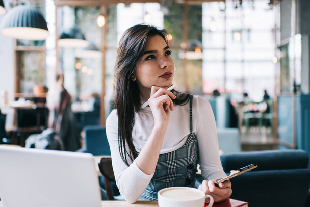 Wistful female freelancer in casual clothes with hand at chin looking away while sitting at table in cafe with clipboard - Foto, Imagen