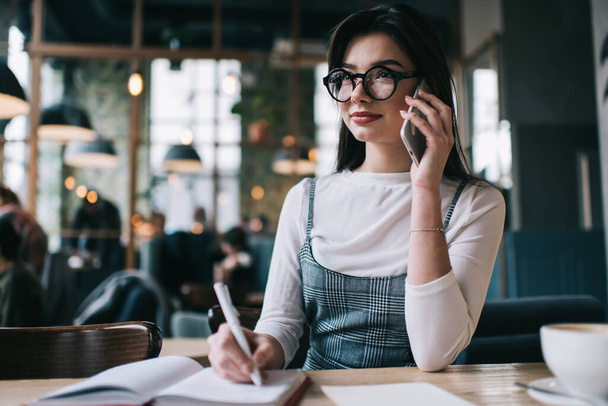 Ικανοποιημένη γυναίκα ελεύθερος επαγγελματίας κοιτάζοντας μακριά, ενώ κάθεται στο καφέ κρατώντας σημειώσεις στο σημειωματάριο και έχοντας συνομιλία στο κινητό τηλέφωνο, ενώ εργάζονται - Φωτογραφία, εικόνα