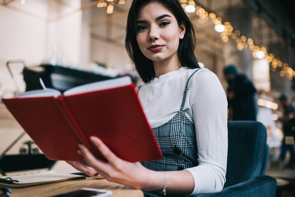 Crop wistful female student in stylish outfit looking at camera while reading notes in notebook and doing task for university - Фото, изображение