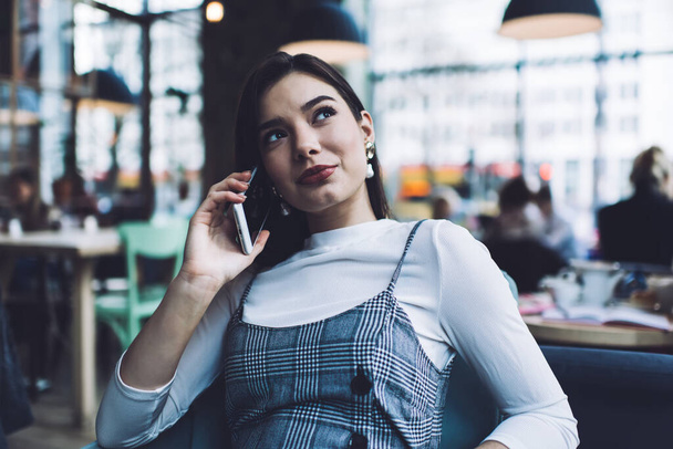 Thoughtful female with dark hair and elegant wear having phone call looking up while sitting in cafeteria on blurred background - Фото, изображение