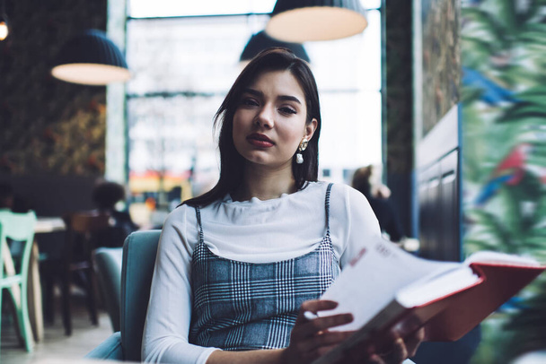 Calm young woman with dark hair in trendy outfit sitting with notebook in hands and thoughtfully looking at camera in cafe in daytime - Фото, изображение