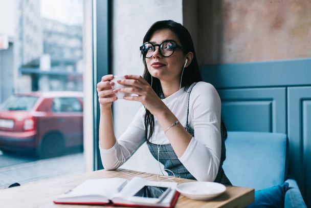 Positive lady in casual wear and eyeglasses looking away and smiling while drinking coffee in cafe and listening to music with earphones - Φωτογραφία, εικόνα